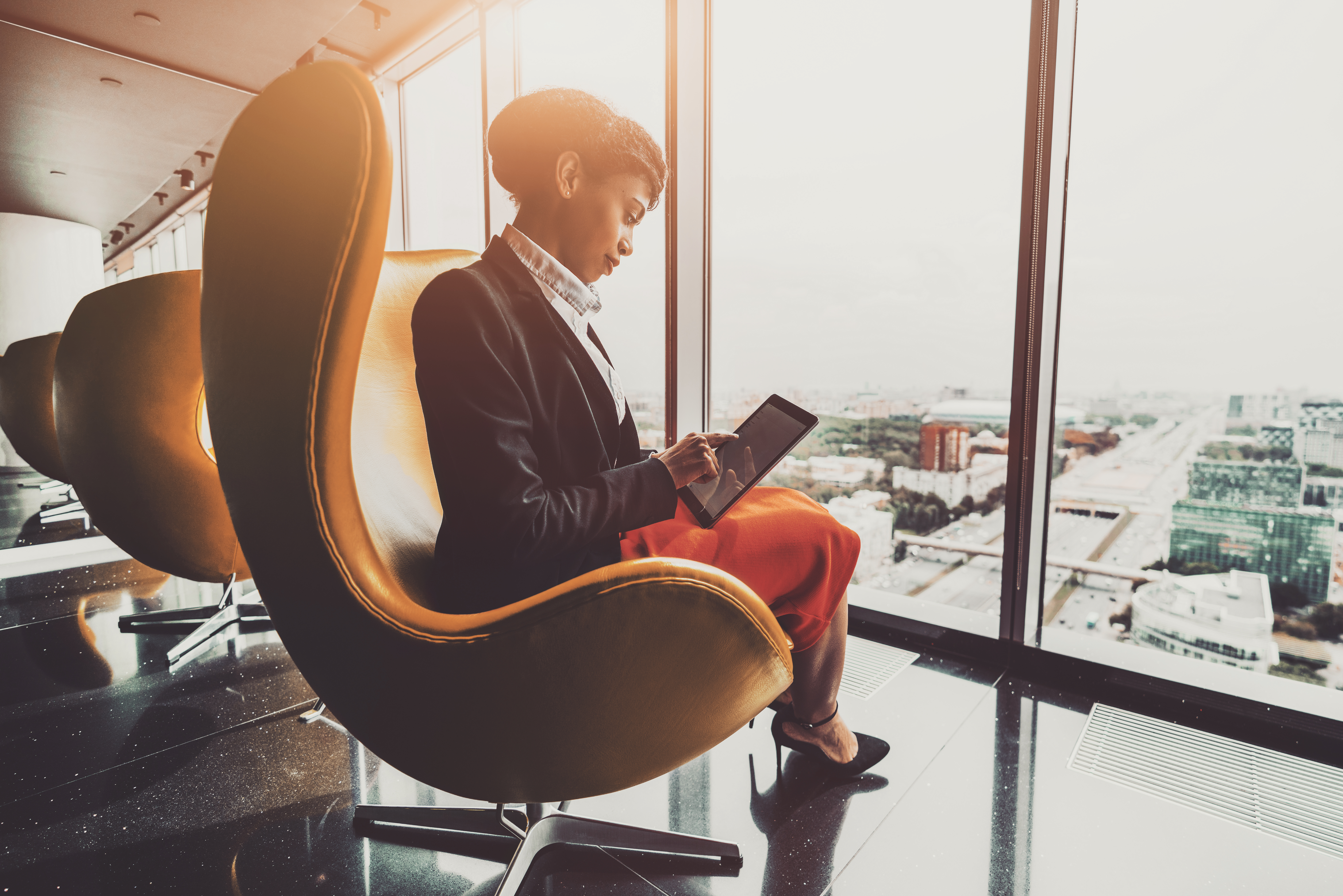 Side view of a dazzling young African-American woman entrepreneur sitting with a digital tablet on a yellow curved armchair indoors of a luxurious office room on a top floor of a business skyscraper