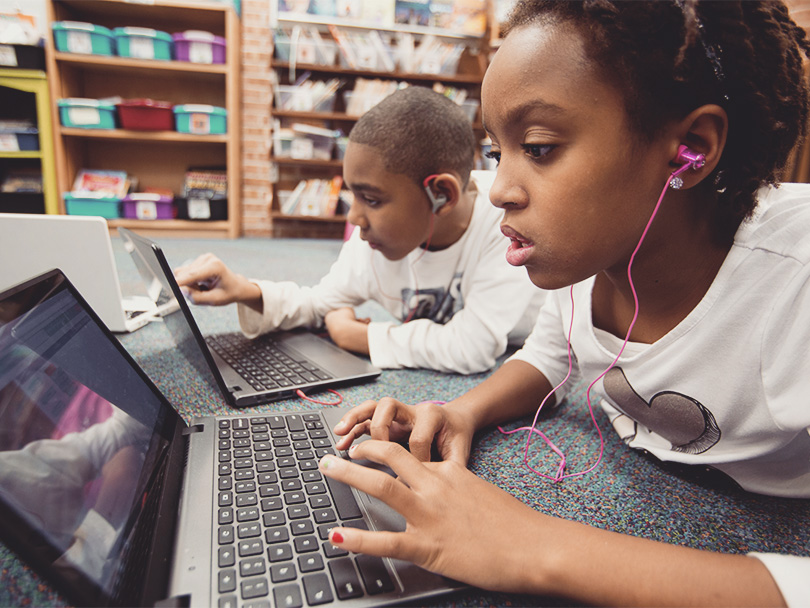 Two fourth grade student work on their laptops side by side sprawled on the classroom floor